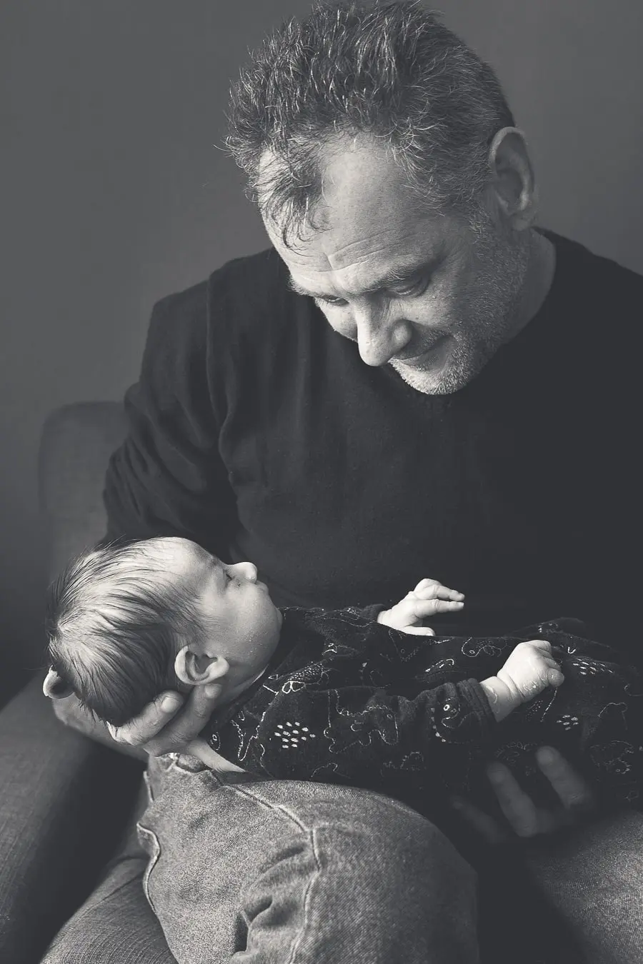Un jeune grand-père regarde avec fierté son petit-fils nouveau-né qu’il tient tendrement dans ses bras. Une photo en noir et blanc capturant un moment intime et émouvant entre deux générations.