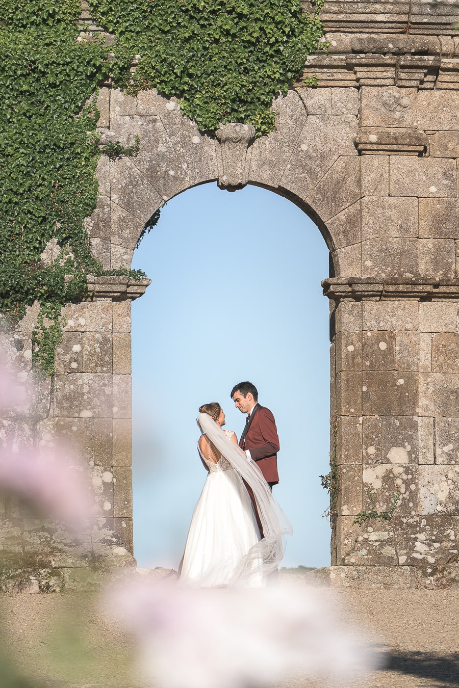 Un couple de jeunes mariés pose sous une arche en pierre recouverte de lierre, lors d'une séance photo de mariage en Bretagne. La mariée porte une robe blanche avec un long voile, et le marié un costume élégant. Un cadre romantique et historique parfait pour une photo de mariage naturelle et intemporelle.