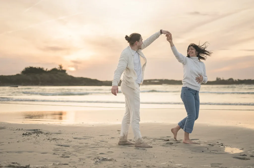 Un couple de fiancés danse pieds nus sur le sable doré d’une plage du Finistère, baigné par la lumière dorée du coucher de soleil.