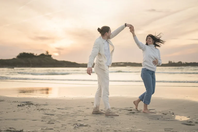 Un couple de fiancés danse pieds nus sur le sable doré d’une plage du Finistère, baigné par la lumière dorée du coucher de soleil.